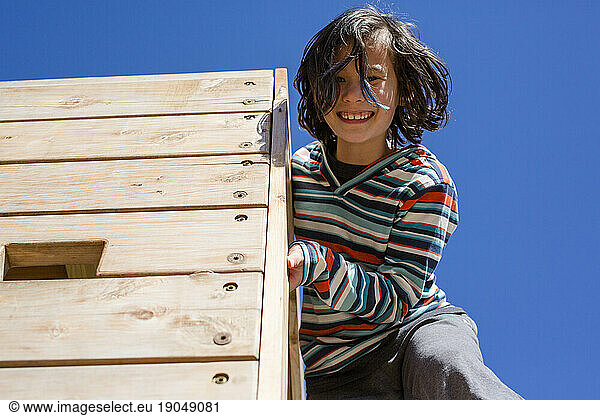 A happy child leans down from a high wooden wall against blue sky