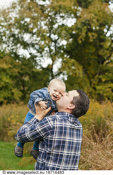 A happy father raises his laughing baby boy up to for a kiss in a park