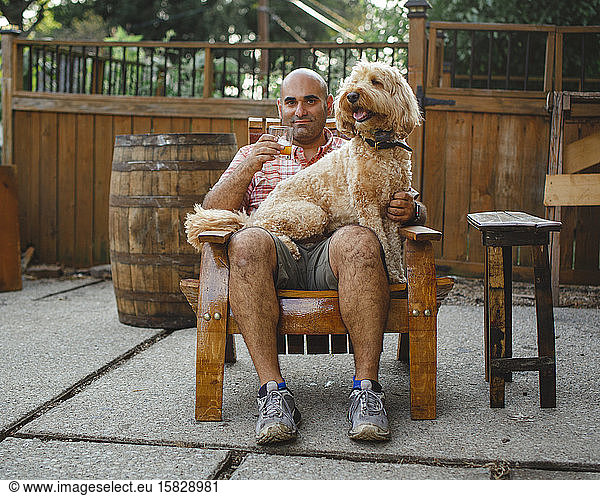 A happy man sits with glass of bourbon and dog on lap on wooden chair