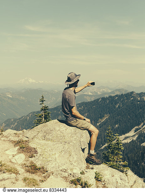 A hiker on a mountain summit  holding a smart phone  at the top of Surprise Mountain  in the Alpine Lakes Wilderness  in Mount Baker-Snoqualmie National Forest.
