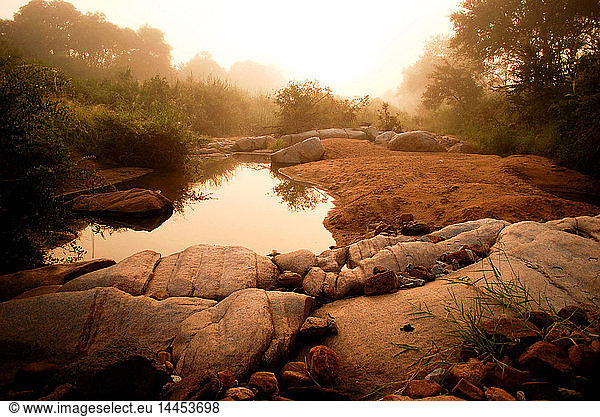 A landscape  a waterhole surrounded by boulders and sand at dusk  with mist in the background