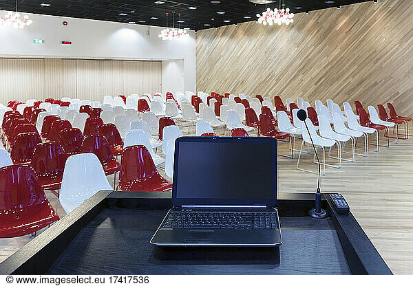 A laptop computer at a podium and rows of chairs in a large room