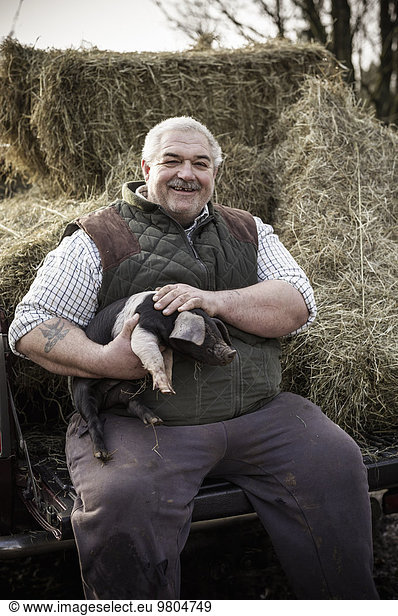 A large man  farmer in a waistcoat and working clothes  holding a piglet and smiling