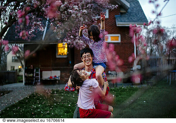 A laughing father carries his two laughing children in yard at dusk