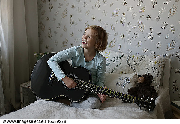 A little blonde plays the guitar and sings in a bright room.