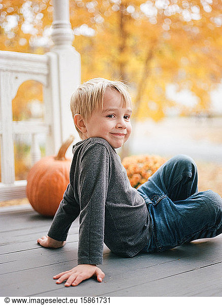 A little boy sitting on a porch by orange maple trees and pumpkins