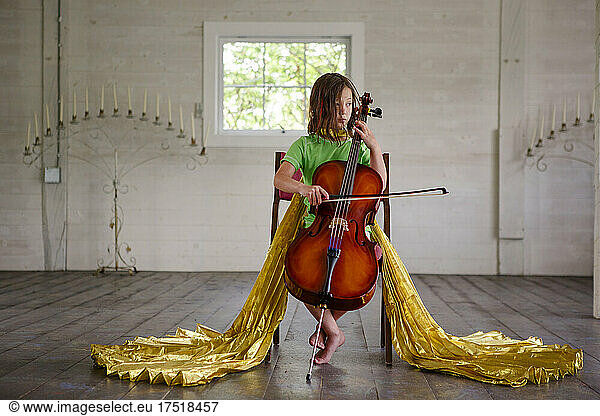 A little girl in long golden wings plays cello in an empty barn