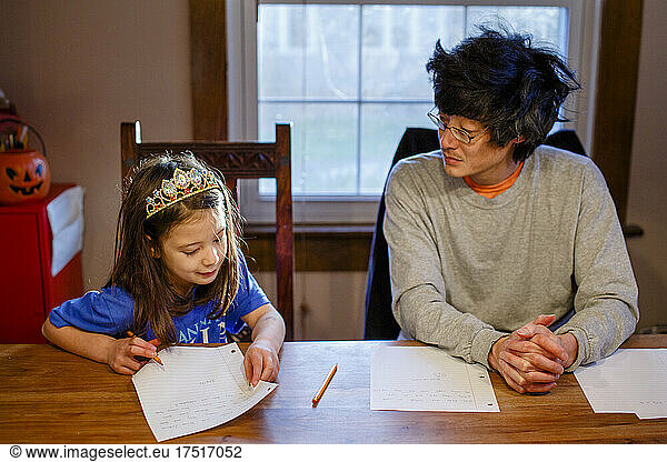 A little girl in princess crown does homework at table with father