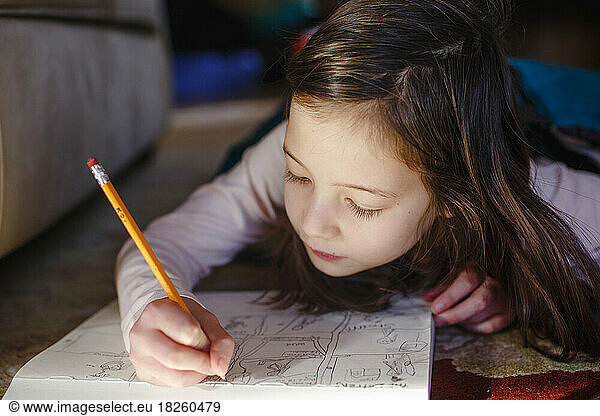 A little girl lays on floor carefully drawing in a sketchpad