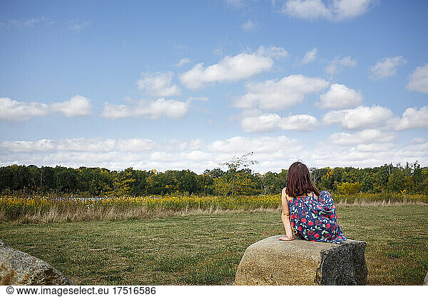 A little girl sits by self on large rock looking out at prairie