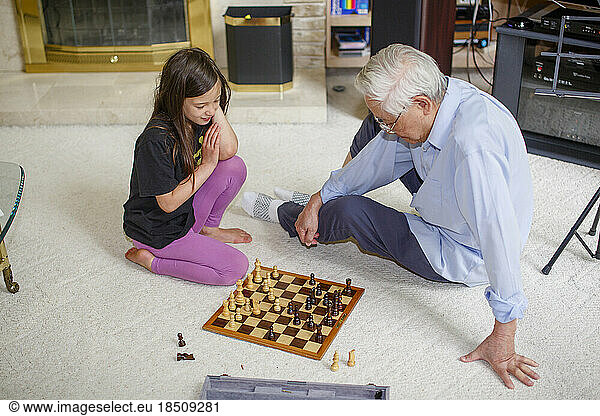 A little girl sits on floor with grandfather playing chess