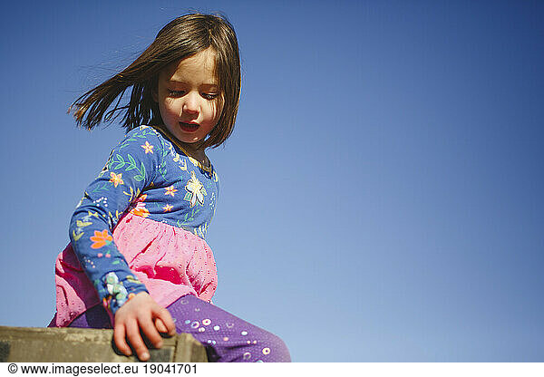 A little girl sits peacefully on a box against blue sky wind in hair