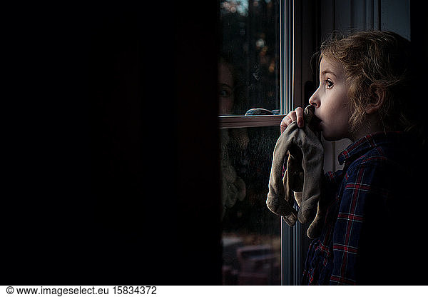A little girl sucking her thumb looks out her front door.