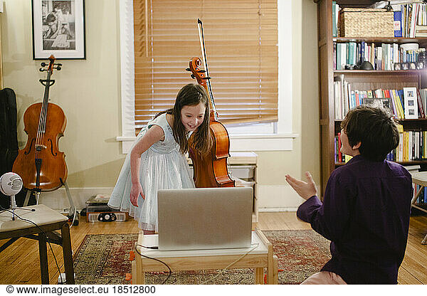 A little girl with cello bows in front of computer while brother claps