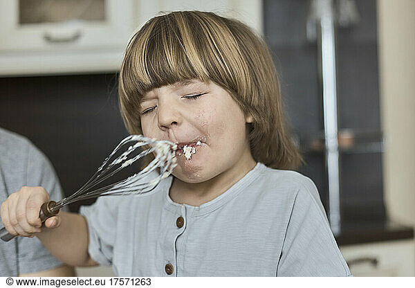 A long-haired boy eats dough off a whisk