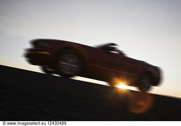A low angle blurred view of a convertible sports car silhouetted on the road at sunset.