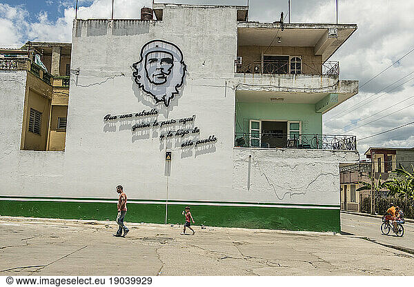 A man and small boy walk under a Graphic of Che Guevara on the Wall of an Apartment House with Propaganda Quote. Santa Clara  Villa Clara  Cuba