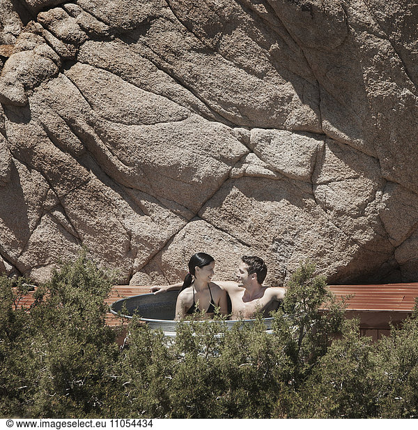 A man and woman on the terrace of an eco home  a low impact house in the desert landscape  in a sunken hot tub.