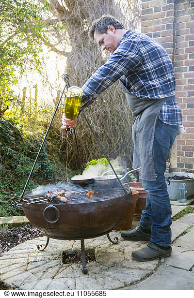 A man cooking roasting game birds over an open fire pouring olive oil from a bottle on the meat.