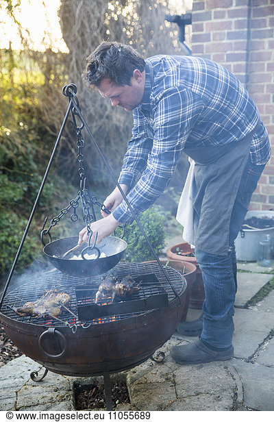 A man cooking vegetables in a pan suspended from a trivet over a glowing fire.