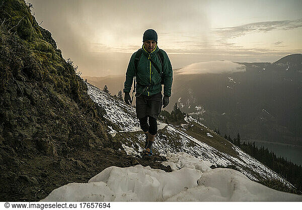 A man hikes up a mountain trail with snow and clouds