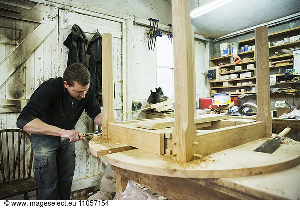 A man in a carpentry workshop  working on the edge of a new wooden table using a hammer.