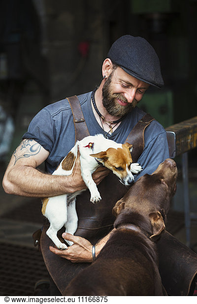 A man in a leather apron playing with two dogs in a workshop.