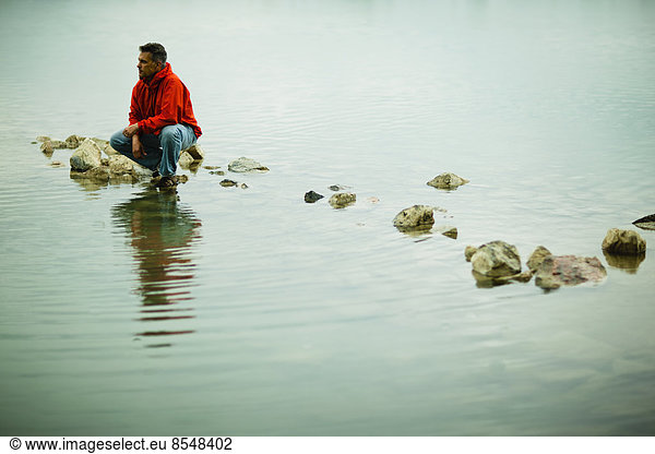 A man in a red jacket balanced in a contemplative pose on a stepping stone or rock in shallow water.