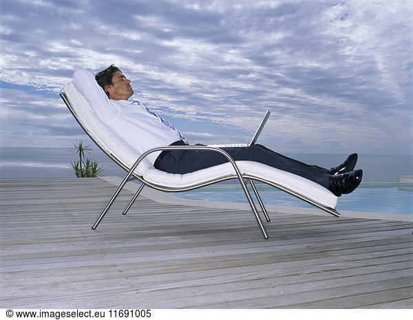A man in a white shirt and black trousers in a sun lounger chair with a laptop on his lap.