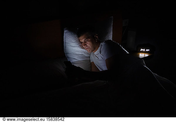 A man is lying in bed in total darkness looking at his smart phone