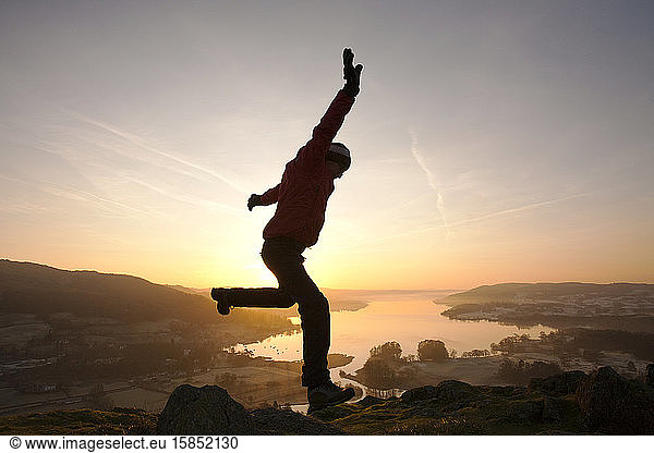 A man jumping for joy at sunrise over Lake Windermere from Todd Crag above Ambleside  Lake District  UK.