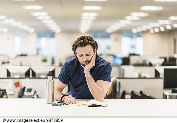 A man leaning on his elbow and looking at an open book on an office desk.