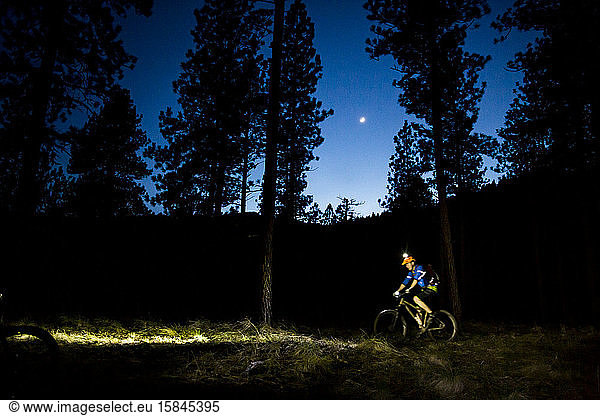 A man mountain bikes under a twilight sky with the moon