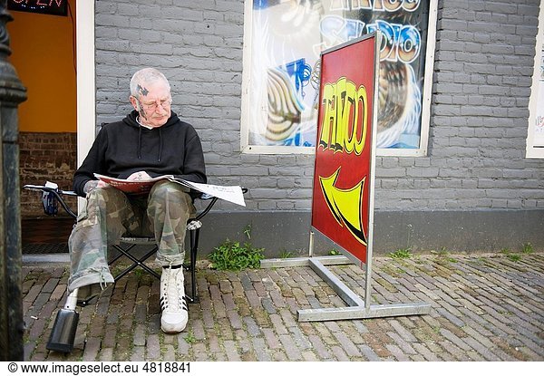 A man  owning a tatooshop  is reading a newspaper outside his shop
