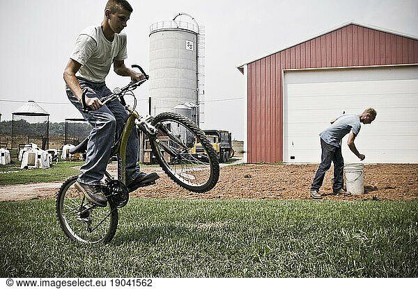 A man prepping the soil to plant crops in the back yard of the farm house while a boy rides his bike in the foreground.