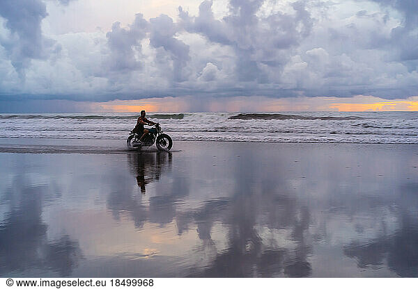 A man rides a motorbike along the beach at sunset.