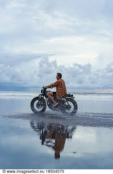 A man rides a motorbike along the beach at sunset.