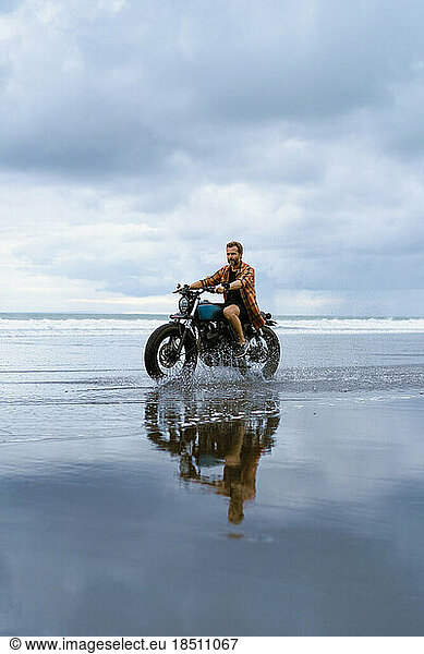 A man rides a motorbike along the beach at sunset.