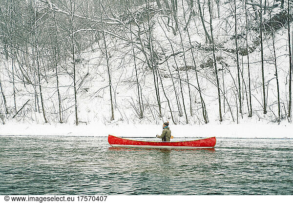 A man riding a red Canadian canoe on the snowy winter river.