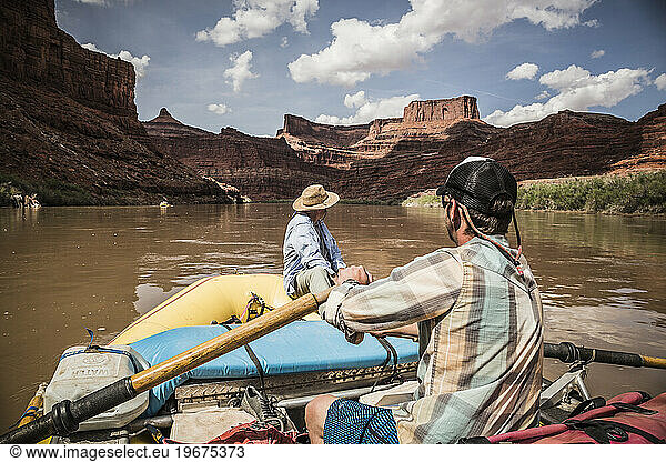 A man rows a raft down a river in a desert landscape.