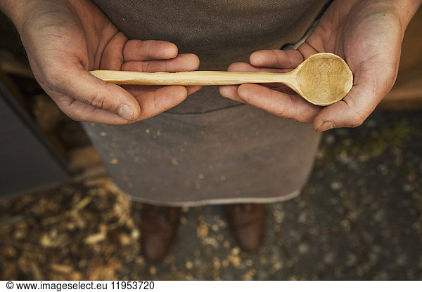 A man's hands holding a hand carved wooden spoon with a long tapering handle and smooth round bowl end.