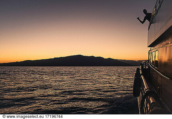 A man's silhouette shows shaka on sunset cruise with Maui in distance