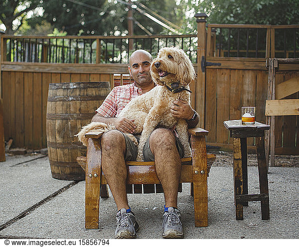 A man sits on a handmade wooden chair with a happy dog on his lap