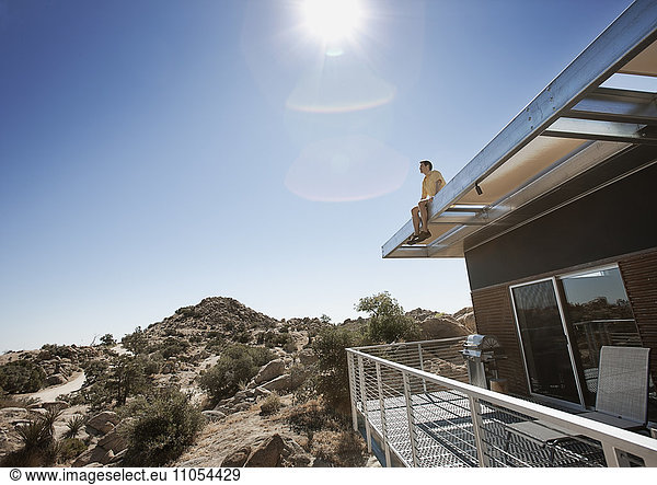 A man sitting on the roof overhang of an eco home in the desert landscape.