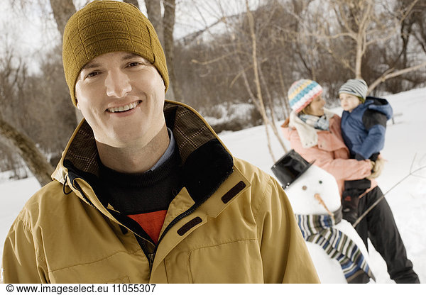 A man smiling in the foreground  and a woman holding a child beside a snowman.