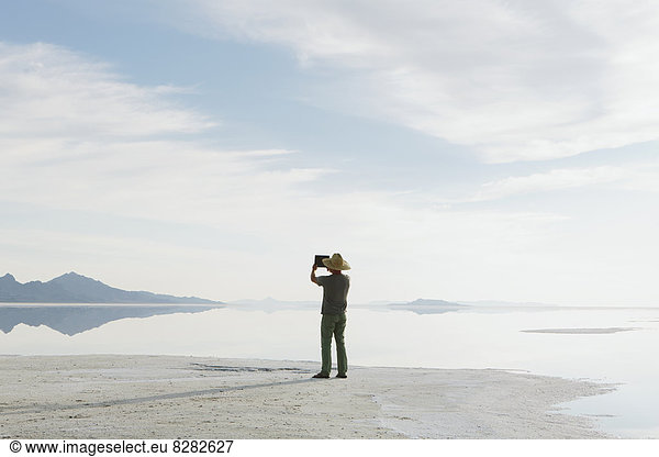 A Man Standing At Edge Of The Flooded Bonneville Salt Flats At Dusk  Taking A Photograph With A Tablet Device.