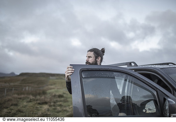 A man standing by an open car door  looking around him  under a cloudy wintery sky.