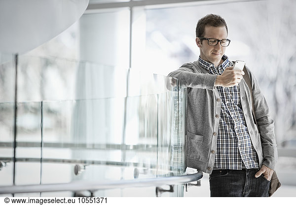 A man standing leaning against a glass panel checking his smart phone.