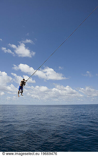 A man swings from a rope over the ocean against a blue sky with white clouds in Costa Rica.
