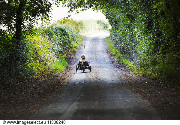 A man using a recumbent three wheeler cycle on a shady country road.